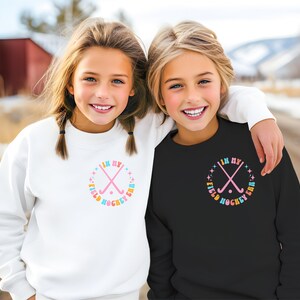 K&ouml;nnte beinhalten: Zwei M&auml;dchen tragen wei&szlig;e und schwarze Sweatshirts mit einer Grafik von gekreuzten Feldhockeyschl&auml;gern und dem Text "In My Field Hockey Era" in einem Regenbogen-Farbschema.
