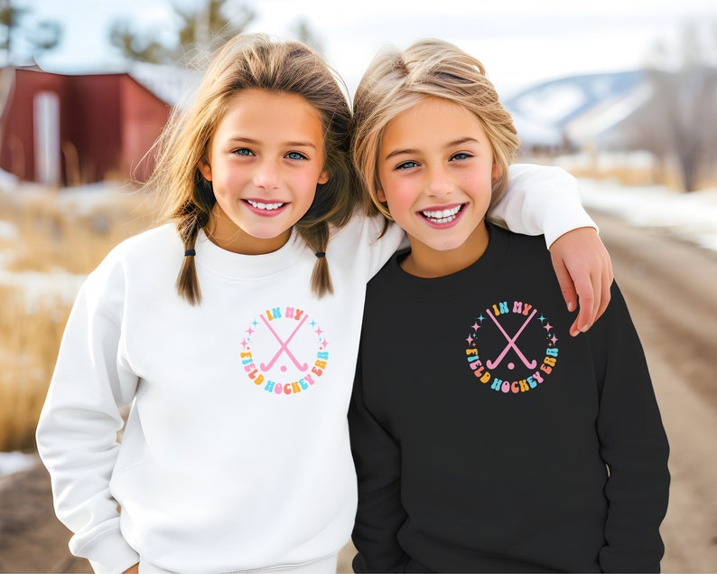K&ouml;nnte beinhalten: Zwei M&auml;dchen tragen wei&szlig;e und schwarze Sweatshirts mit einer Grafik von gekreuzten Feldhockeyschl&auml;gern und dem Text "In My Field Hockey Era" in einem Regenbogen-Farbschema.