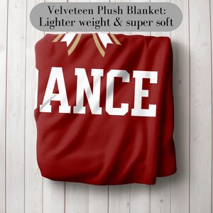 K&ouml;nnte beinhalten: Eine rote Velours-Pl&uuml;schdecke mit dem Wort "ANCE" in gro&szlig;en wei&szlig;en Buchstaben. Die Decke hat goldene Akzente und den Text "Velveteen Plush Blanket: Lighter weight & super soft" in einem grauen Oval.