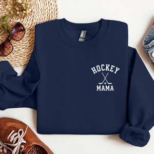 Op de afbeelding: Navyblauwe trui met witte tekst die "Hockey Mama" zegt en een afbeelding van twee gekruiste hockeysticks met een puck.