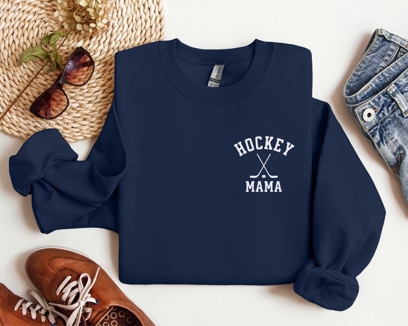 Op de afbeelding: Navyblauwe trui met witte tekst die "Hockey Mama" zegt en een afbeelding van twee gekruiste hockeysticks met een puck.