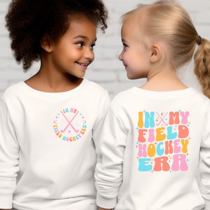 K&ouml;nnte beinhalten: Zwei Kinder tragen wei&szlig;e Langarmshirts mit einem Feldhockey-Design. Das Shirt auf der linken Seite hat ein pinkfarbenes, blaues und orangefarbenes Design mit gekreuzten Feldhockeyschl&auml;gern und dem Text "In My Field Hockey Era". Das Shirt auf der rechten Seite hat den gleichen Text in einer gr&ouml;&szlig;eren Schriftart mit einem pinkfarbenen, blauen, orangen und gelben Farbschema.