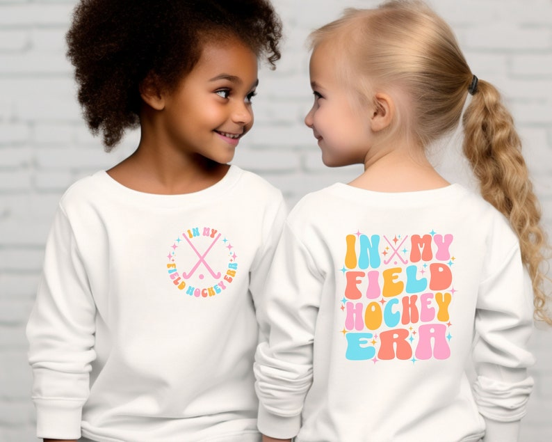 K&ouml;nnte beinhalten: Zwei Kinder tragen wei&szlig;e Langarmshirts mit einem Feldhockey-Design. Das Shirt auf der linken Seite hat ein pinkfarbenes, blaues und orangefarbenes Design mit gekreuzten Feldhockeyschl&auml;gern und dem Text "In My Field Hockey Era". Das Shirt auf der rechten Seite hat den gleichen Text in einer gr&ouml;&szlig;eren Schriftart mit einem pinkfarbenen, blauen, orangen und gelben Farbschema.