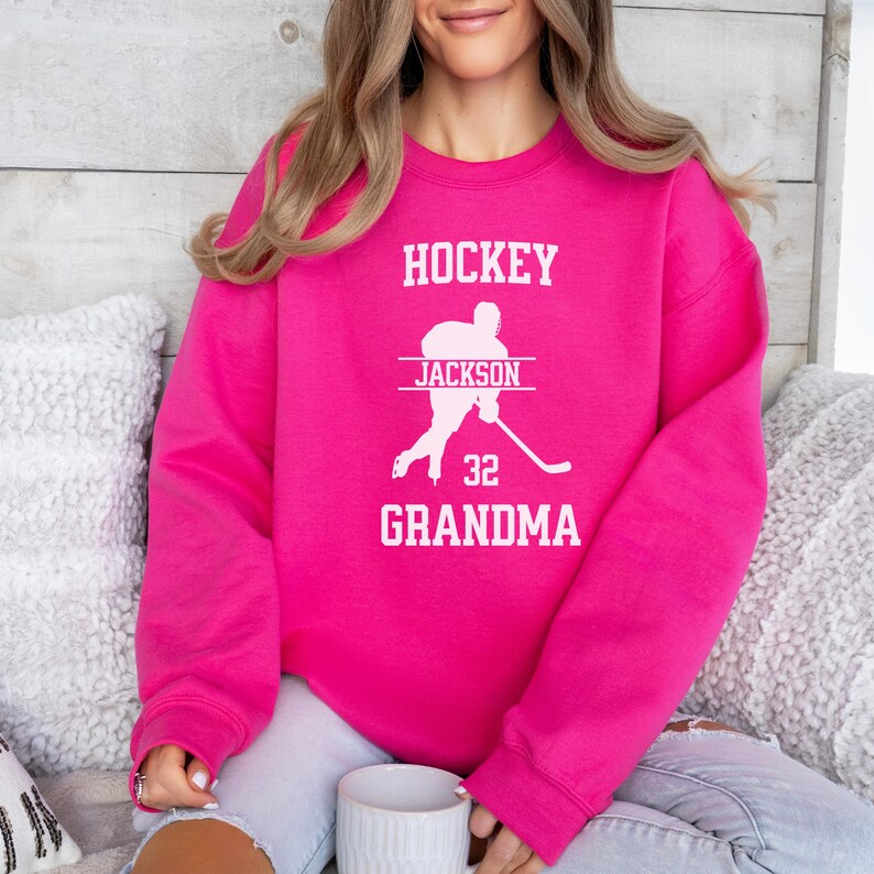 May include: Pink sweatshirt with the text "HOCKEY" and "GRANDMA" in white. A white silhouette of a hockey player with the name "JACKSON" and the number "32" is printed on the sweatshirt.