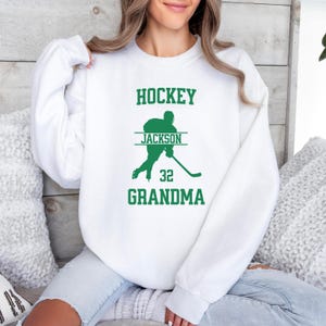 May include: White sweatshirt with green text that reads "HOCKEY" above a silhouette of a hockey player. The text "JACKSON" is above the player's number "32" and the text "GRANDMA" is below the player.