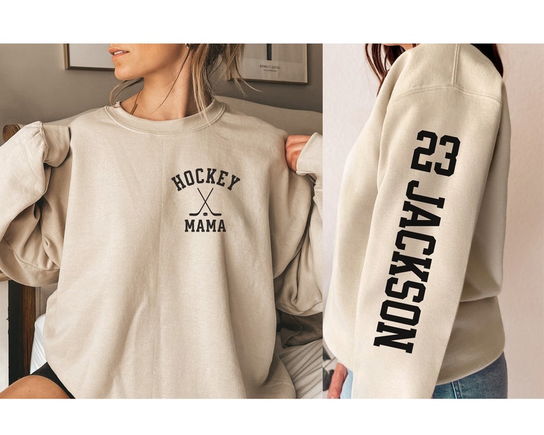Gildan brand, Sand colored crewneck sweatshirt with Hockey Mama written on the left front crest, along with a customizable sleeve print with the example number 23 on the left shoulder and the example name running the length of the sleeve.