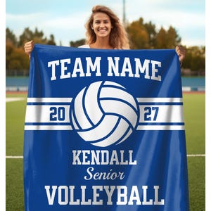 May include: A blue rectangular blanket featuring a white volleyball graphic in the center. The text reads TEAM NAME, 20, 27, KENDALL, Senior, and VOLLEYBALL in white lettering. A horizontal white stripe runs across the middle behind the volleyball.