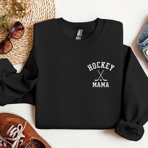 Op de afbeelding: Zwart sweatshirt met witte tekst die "Hockey Mama" zegt en een afbeelding van twee gekruiste hockeysticks en een puck.