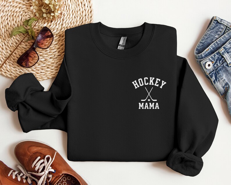 Op de afbeelding: Zwart sweatshirt met witte tekst die "Hockey Mama" zegt en een afbeelding van twee gekruiste hockeysticks en een puck.