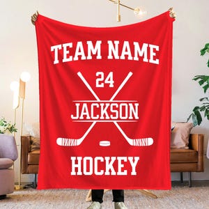 May include: A bright red rectangular fleece blanket featuring white printed text and graphics. The design includes the words TEAM NAME, JACKSON, and HOCKEY, along with a large graphic of two crossed hockey sticks, a puck, and the number 24.