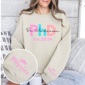 May include: A light beige sweatshirt with the text "Dr. Anderson PhD Est. 2024" in pink, blue, and black lettering. The sleeves have the word "Physics" in pink lettering with a graduation cap icon.