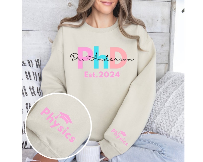 May include: A light beige sweatshirt with the text "Dr. Anderson PhD Est. 2024" in pink, blue, and black lettering. The sleeves have the word "Physics" in pink lettering with a graduation cap icon.