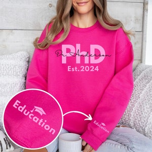 May include: Pink sweatshirt with the text "Dr. Anderson PhD Est. 2024" and a graduation cap graphic. The sweatshirt has the word "Education" printed on the sleeve.