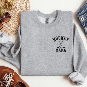 Op de afbeelding: Grijs sweatshirt met de tekst "Hockey Mama" en een afbeelding van gekruiste hockeysticks en een puck.