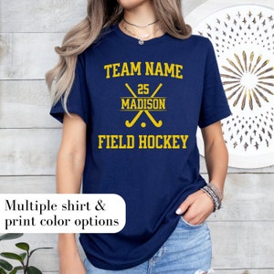 May include: Navy blue t-shirt with yellow text. The shirt features the words "TEAM NAME" above crossed field hockey sticks and the name "MADISON" with the number "25". Below the sticks, it says "FIELD HOCKEY". The shirt also says "Multiple shirt & print color options".