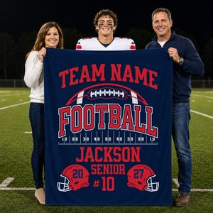 May include: A navy blue rectangular fleece blanket featuring a red and white graphic design with the text TEAM NAME, FOOTBALL, JACKSON, SENIOR, and #10. The design includes a football illustration, yard lines, and two red football helmets.