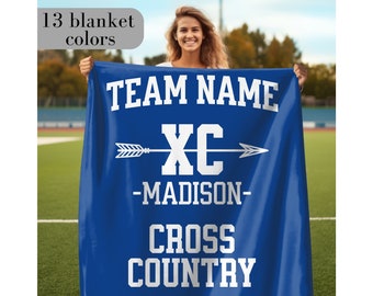 May include: Blue fleece blanket with white text that reads "TEAM NAME XC -MADISON- CROSS COUNTRY". An arrow with a white outline points to the right of the text "XC".