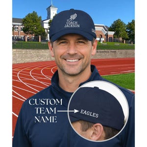 May include: A navy blue baseball cap with the text "COACH JACKSON" and a wing design. The cap also features the word "EAGLES" on the side. The image shows a man wearing the cap in front of a track and field.