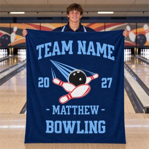 May include: A dark blue rectangular blanket featuring light blue text that reads TEAM NAME, MATTHEW, and BOWLING. The design includes a graphic of a black bowling ball hitting white bowling pins with red stripes, flanked by the numbers 20 and 27.
