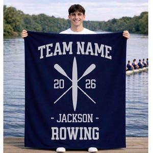 May include: Navy blue blanket with white text and a rowing theme. The blanket features the words "TEAM NAME" at the top, crossed oars, the numbers "20" and "26", and the words "- JACKSON - ROWING" at the bottom. A person is holding the blanket.