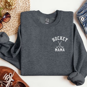 Op de afbeelding: Een donkergrijs sweatshirt met een witte afbeelding waarop "HOCKEY MAMA" staat met twee gekruiste hockeysticks en een puck.