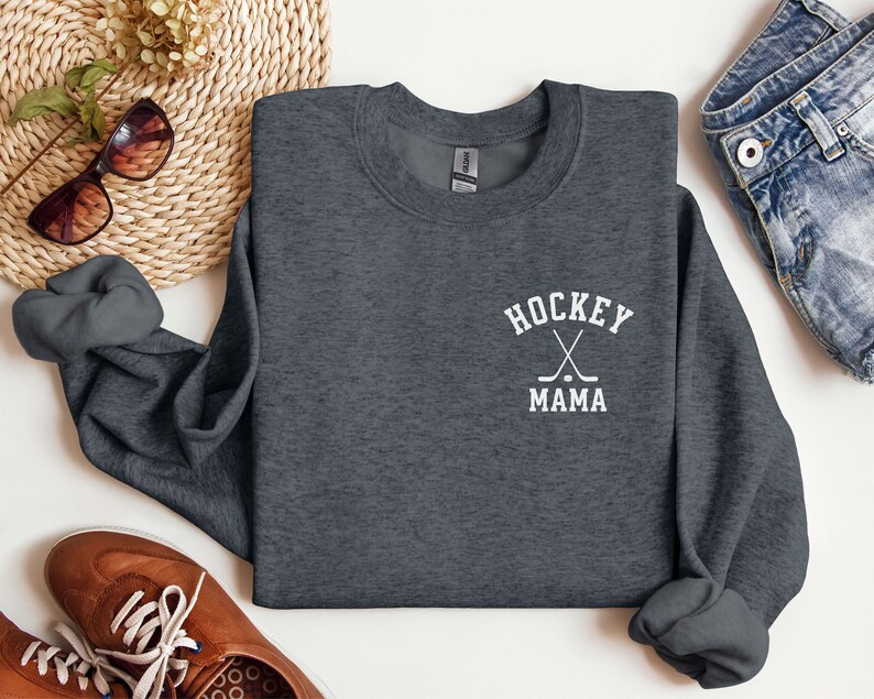 Op de afbeelding: Een donkergrijs sweatshirt met een witte afbeelding waarop "HOCKEY MAMA" staat met twee gekruiste hockeysticks en een puck.