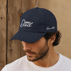 May include: Navy blue baseball cap with the words "Bonus Dad" embroidered in white script on the front. The text "Since 2023" is embroidered on the side. The cap is made of a soft material.