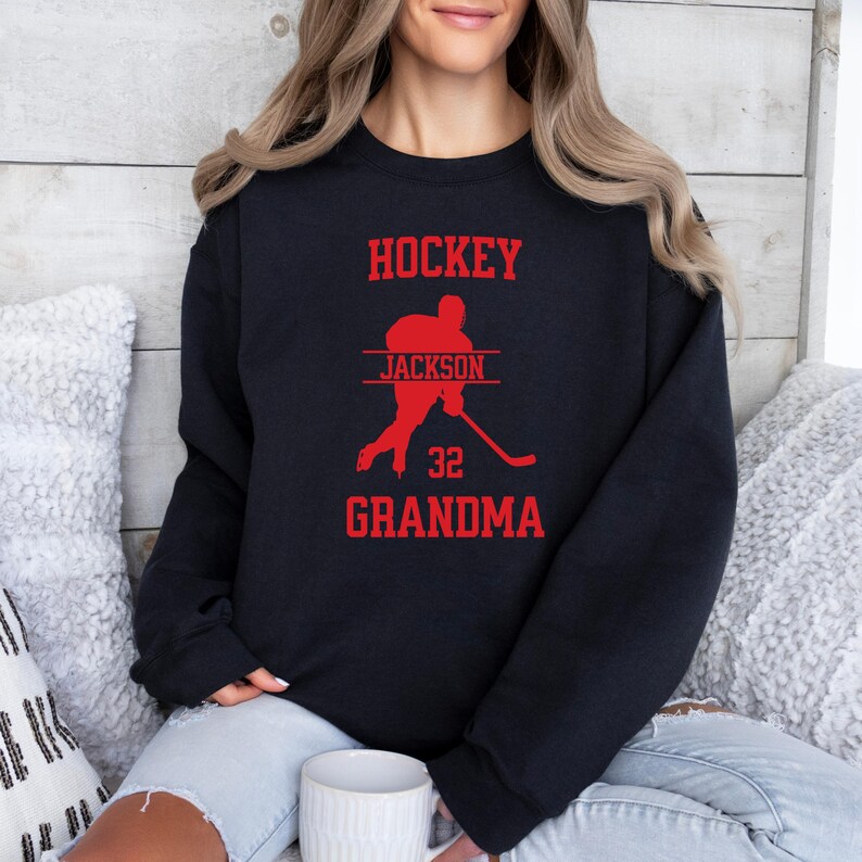 May include: Black sweatshirt with red text that says "Hockey" and "Grandma". A red silhouette of a hockey player with the name "Jackson" and the number "32" is in the center of the sweatshirt.