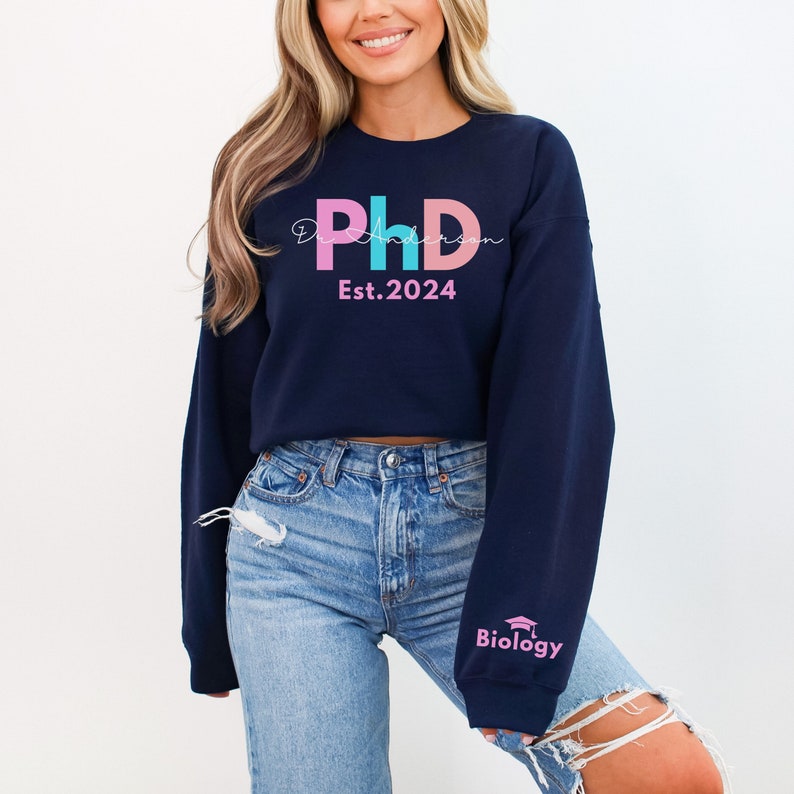 May include: A navy blue cropped sweatshirt with the text "PhD" in pink, blue, and light pink letters. The text "Est. 2024" is below the letters. The left sleeve has the text "Biology" in pink letters with a graduation cap icon.