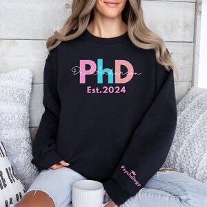 May include: Black sweatshirt with a pink, blue, and pink "PhD" graphic with the text "Dr. Anderson Est. 2024" and a small graphic of a graduation cap with the text "Psychology" on the sleeve.