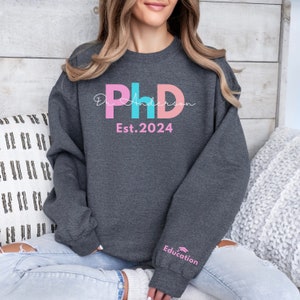 May include: A dark gray sweatshirt with a pink, blue, and white graphic that says "Dr. Anderson PhD Est. 2024". The sweatshirt has a pink and white graphic on the sleeve that says "Education".