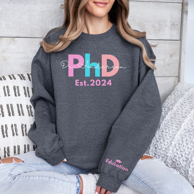 May include: A dark gray sweatshirt with a pink, blue, and white graphic that says "Dr. Anderson PhD Est. 2024". The sweatshirt has a pink and white graphic on the sleeve that says "Education".