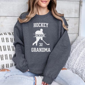 May include: A dark gray sweatshirt with the text "HOCKEY" in white, followed by a silhouette of a hockey player with the name "JACKSON" and the number "32" printed below. The text "GRANDMA" is printed below the player.