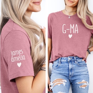 May include: Two pink t-shirts with white text. One shirt says "G-MA" with a small heart. The other shirt has "james amelia" and a heart on the sleeve.