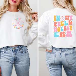 K&ouml;nnte beinhalten: Wei&szlig;er Pullover mit einer pinkfarbenen, blauen und gelben Grafik, auf der hinten "In My Field Hockey Era" steht. Die Vorderseite des Pullovers hat eine kleinere Grafik mit zwei gekreuzten Hockeyschl&auml;gern in einem Kreis.
