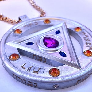 Puede incluir: Collar con colgante de plata con un diseño triangular y piedras preciosas. El triángulo tiene una gran piedra preciosa púrpura en el centro y piedras preciosas azules más pequeñas en los puntos. El colgante está rodeado por un círculo con escritura hebrea y piedras preciosas naranjas más pequeñas.