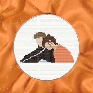 Puede incluir: Bordado de punto de cruz de dos personas abrazándose, una lleva una chaqueta negra y la otra una camisa naranja.