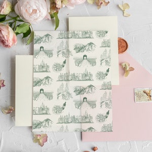 Op de afbeelding: Een set huwelijksuitnodigingen met een roze envelop, een cr&egrave;me envelop en een cr&egrave;me kaart met een groen-wit patroon van stadsgezichten en bergen. De kaart heeft een zegel van was.
