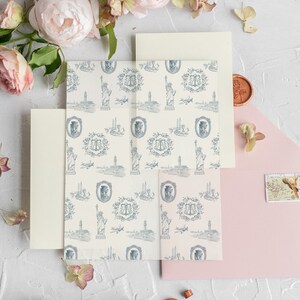 Op de afbeelding: Een set huwelijksuitnodigingen met een lichtroze envelop, een cr&egrave;me envelop en een cr&egrave;me kaart met een blauw-wit patroon van het Vrijheidsbeeld, vliegtuigen en een monogram.