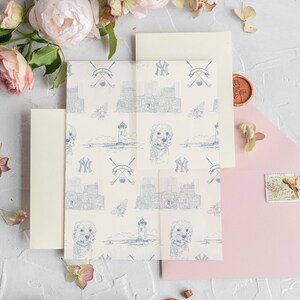 Op de afbeelding: Een set huwelijksuitnodigingen met een witte achtergrond en een blauw-wit patroon. Het patroon bevat een vuurtoren, een hond en de letters "NY". De uitnodigingen liggen op een wit oppervlak met roze en witte bloemen.