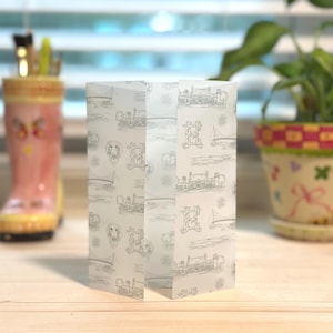 May include: A folded, translucent paper gift bag with a repeating pattern of vintage illustrations, including trains, sailboats, and dogs. The bag is standing upright on a wooden surface, with a blurred background.