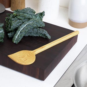 May include: A light-colored wooden spatula with a wide, leaf-shaped head and a long, slender handle, resting on a dark brown, thick, rectangular wooden cutting board next to a bunch of dark green, curly kale leaves.