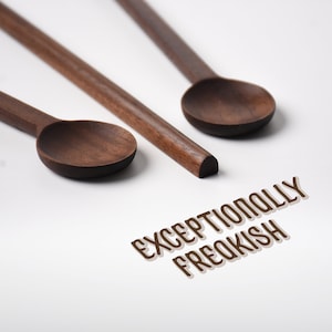 May include: Two dark brown wooden spoons with long, slender handles and rounded bowls. The text "EXCEPTIONALLY FREAKISH" is printed in a brown, sans-serif font with a drop shadow effect below the spoons on a plain white surface.