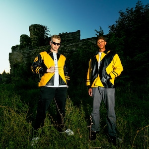 May include: Two people wearing black and yellow jackets with black trousers and sunglasses. The jackets feature a distinctive design with black and yellow panels and the number 8 on the left shoulder. A stone structure and green foliage are in the background.