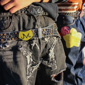 May include: Two people wearing jeans with embellished belts. The belts have black and silver rhinestones and a large metal buckle with a yellow "M" logo. One pair of jeans is ripped and distressed.