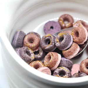 K&ouml;nnte beinhalten: Eine wei&szlig;e Sch&uuml;ssel gef&uuml;llt mit lila und rosa Donut-f&ouml;rmigem M&uuml;sli. Ein silberner L&ouml;ffel befindet sich in der Sch&uuml;ssel und sch&ouml;pft etwas M&uuml;sli heraus.