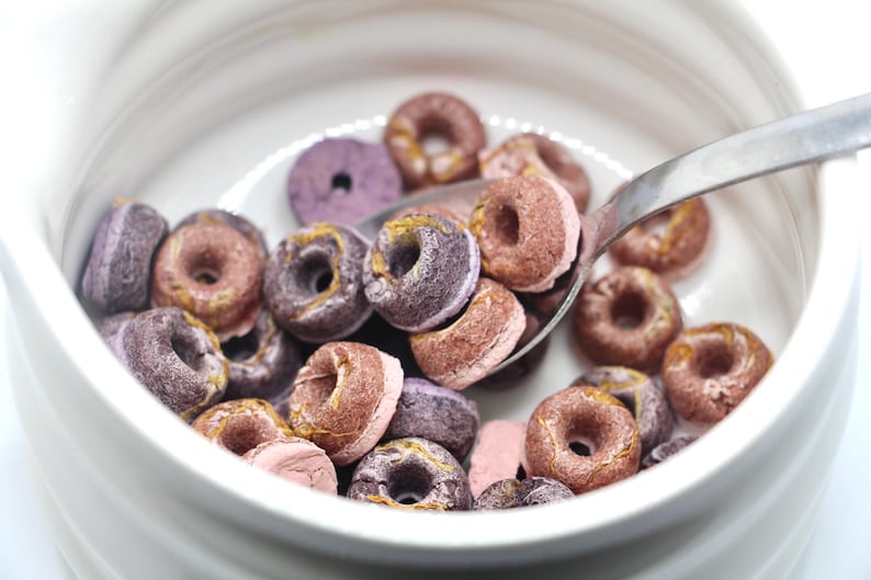 K&ouml;nnte beinhalten: Eine wei&szlig;e Sch&uuml;ssel gef&uuml;llt mit lila und rosa Donut-f&ouml;rmigem M&uuml;sli. Ein silberner L&ouml;ffel befindet sich in der Sch&uuml;ssel und sch&ouml;pft etwas M&uuml;sli heraus.