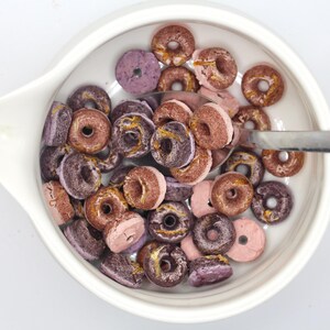 K&ouml;nnte beinhalten: Eine wei&szlig;e Sch&uuml;ssel gef&uuml;llt mit rosafarbenem und violettem, glasiertem Donut-f&ouml;rmigem M&uuml;sli. Ein silberner L&ouml;ffel befindet sich in der Sch&uuml;ssel.