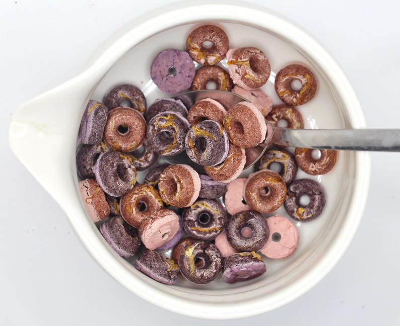 K&ouml;nnte beinhalten: Eine wei&szlig;e Sch&uuml;ssel gef&uuml;llt mit rosafarbenem und violettem, glasiertem Donut-f&ouml;rmigem M&uuml;sli. Ein silberner L&ouml;ffel befindet sich in der Sch&uuml;ssel.