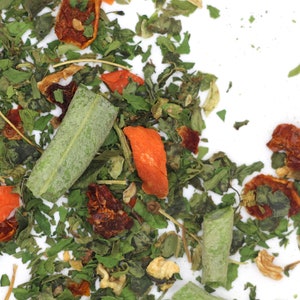 May include: A close-up of a mix of dried herbs and vegetables, including green leafy herbs, orange and red dried fruit pieces, and some larger green pieces.